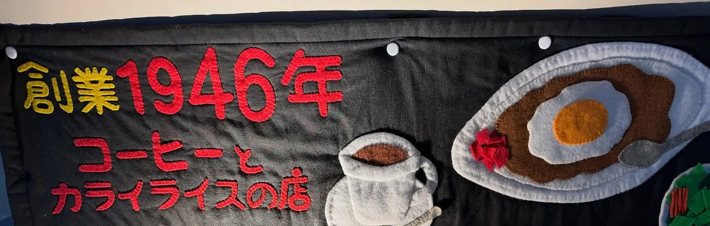 黒い布地に「創業1946年 コーヒーとカライライスの店」と縫い付けた看板。コーヒーカップと目玉焼きのカレーの絵